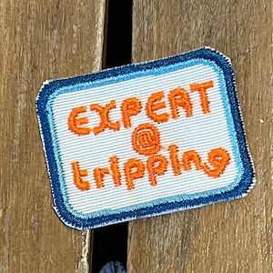 May include: Embroidered patch with the words "EXPERT @ tripping" in orange thread on a white background. The patch has a blue border with a thin light blue outline. The patch is rectangular.