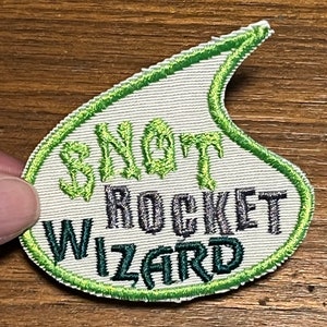 May include: Embroidered patch in a teardrop shape with a green border. The patch features the words "SNOT ROCKET WIZARD" in green and gray thread on a white background. The patch is likely for clothing or accessories.