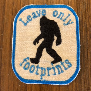May include: A white patch with a blue border features a black silhouette of a Bigfoot walking with the text "Leave only footprints" in blue.