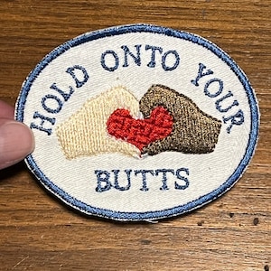 May include: Oval embroidered patch with the text "HOLD ONTO YOUR BUTTS" in blue and white. The patch features two embroidered hands holding a red heart in the center. The hands are beige and brown.