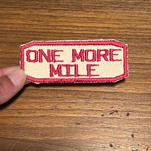 May include: A red and white embroidered patch with the text "ONE MORE MILE" in red.