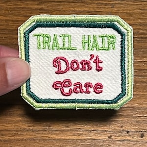 May include: White embroidered patch with green and teal stitching. The patch reads "Trail Hair Don't Care" in green and red lettering.