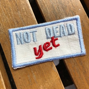 May include: A white fabric patch with blue stitching around the edges. The text "NOT DEAD yet" is embroidered on the patch in blue and red thread.