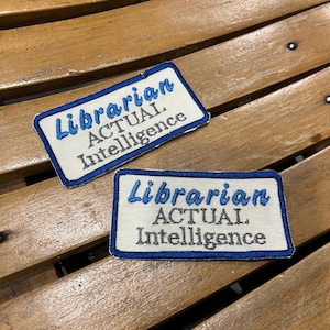 May include: Two rectangular embroidered patches with a white background and a blue border. The text "Librarian" is stitched in blue script, while "ACTUAL Intelligence" is stitched in black block lettering below it on both patches.