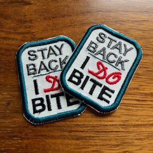 May include: Two rectangular embroidered patches with the text "STAY BACK I DO BITE". The patches have a white background, black and red lettering, and a teal border. The patches are on a wooden surface.