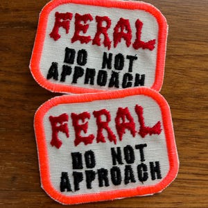 May include: Two rectangular patches with the words "FERAL DO NOT APPROACH" in red and black lettering. The patches have a white background with an orange border.