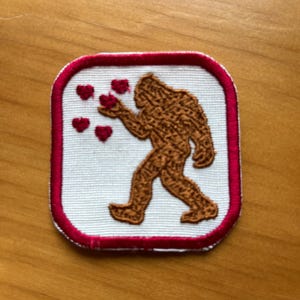 May include: A white embroidered patch with a red border. The patch features a brown Bigfoot figure holding out a hand with five red hearts floating around it.