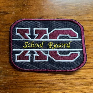 May include: A rectangular patch with a navy blue background and a maroon border. The patch features the letters "XC" in maroon, with "School Record" in yellow script below. The patch is likely for a school sports team.