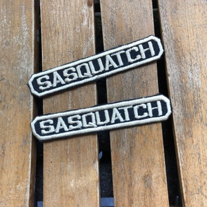 May include: Two black and white embroidered patches with the word "Sasquatch" in white lettering. The patches are on a wooden surface.