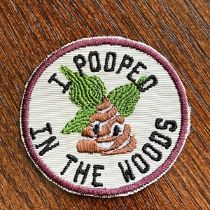 May include: Embroidered patch with the text "I Pooped in the Woods" in black lettering. The patch features a cartoon poop emoji with green leaves and a brown and white color scheme, set against a white background with a maroon border.