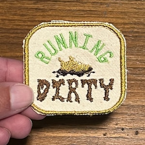 May include: A beige fabric patch with a gold border. The patch has green embroidery that reads "RUNNING DIRTY" with a brown embroidered image of a car in the mud.