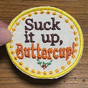 May include: A white embroidered patch with a yellow border and a brown and orange floral design. The text on the patch reads "Suck it up, Buttercup!"