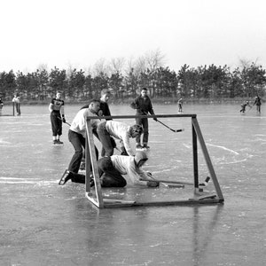 May include: A group of people playing hockey on a frozen lake. One player is on the ground trying to stop the puck from going into the goal. The goal is made of metal and has a net. The players are wearing winter clothing and are having fun.