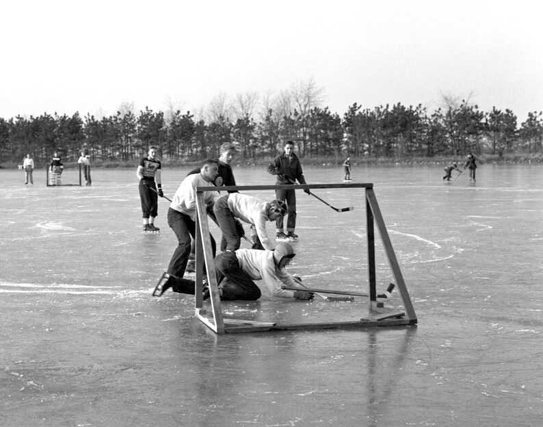 May include: A group of people playing hockey on a frozen lake. One player is on the ground trying to stop the puck from going into the goal. The goal is made of metal and has a net. The players are wearing winter clothing and are having fun.
