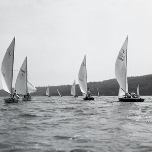 Puede incluir: Imagen en blanco y negro de veleros en un lago. Varios peque&ntilde;os veleros con velas blancas navegan sobre el agua. Las velas tienen n&uacute;meros y un logotipo. El fondo muestra una l&iacute;nea de &aacute;rboles y un cielo nublado.