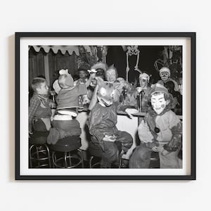 May include: A black and white photograph in a black frame depicting a group of children in various costumes, including animal masks and clown outfits, sitting at a counter. The scene features a skeleton decoration in the background and multiple stools.