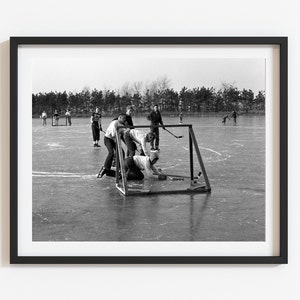 May include: A black and white photo of a group of people playing hockey on a frozen lake. The players are wearing winter clothing and are using hockey sticks to hit a puck. There is a goal in the background.