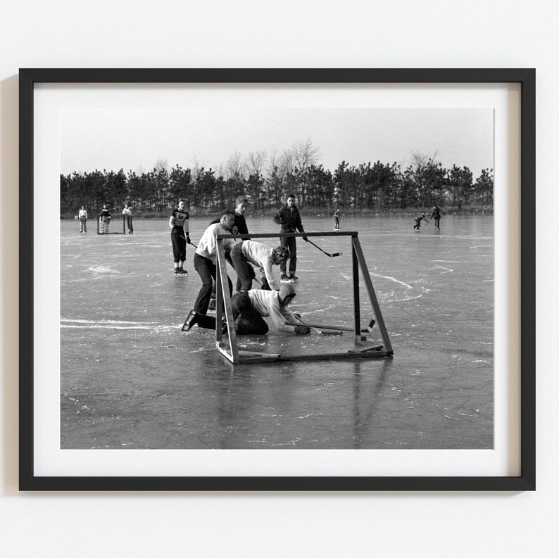 May include: A black and white photo of a group of people playing hockey on a frozen lake. The players are wearing winter clothing and are using hockey sticks to hit a puck. There is a goal in the background.