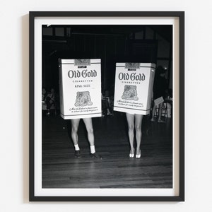 May include: Two people are dressed up in cardboard boxes that look like Old Gold cigarette packs. The boxes are white with black and gold lettering. The text on the boxes reads "Old Gold Cigarettes King Size". The people are standing on a wooden floor.
