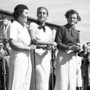 Pode incluir: Tr&ecirc;s mulheres com vestidos brancos e chap&eacute;us est&atilde;o juntas segurando tacos de golfe. Elas est&atilde;o sorrindo e olhando para a c&acirc;mera. As mulheres est&atilde;o em um campo de golfe.