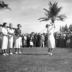 Pode incluir: Tr&ecirc;s mulheres com vestidos e chap&eacute;us brancos est&atilde;o em um campo de golf, observando uma mulher com um vestido e chap&eacute;u brancos balan&ccedil;ando um taco de golfe. A mulher est&aacute; no meio do seu swing e o taco est&aacute; prestes a atingir a bola. H&aacute; muitas pessoas no fundo assistindo ao jogo.