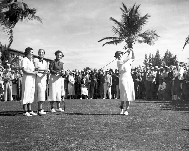 Pode incluir: Tr&ecirc;s mulheres com vestidos e chap&eacute;us brancos est&atilde;o em um campo de golf, observando uma mulher com um vestido e chap&eacute;u brancos balan&ccedil;ando um taco de golfe. A mulher est&aacute; no meio do seu swing e o taco est&aacute; prestes a atingir a bola. H&aacute; muitas pessoas no fundo assistindo ao jogo.