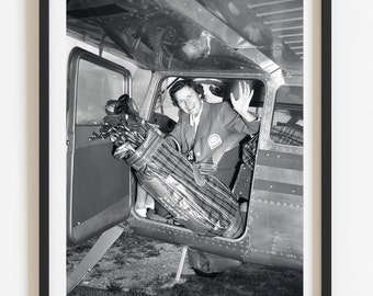 Puede incluir: Una fotografía en blanco y negro de una mujer sentada en la puerta abierta de un pequeño avión. Ella sostiene una bolsa de golf con palos que sobresalen de la parte superior. Ella sonríe y saluda. La foto está enmarcada en un marco negro.