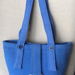 May include: A blue woven tote bag with two long straps and a silver ring on the front. The bag has two blue buttons on the straps.