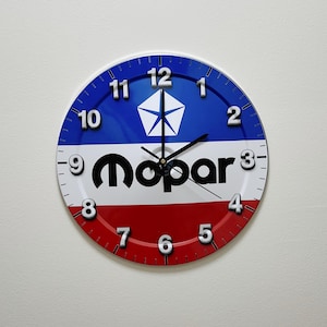 May include: A round wall clock featuring a horizontal tricolor design of blue, white, and red. The center displays the black Mopar logo and a white pentastar emblem. The clock face includes raised silver Arabic numerals and black clock hands.