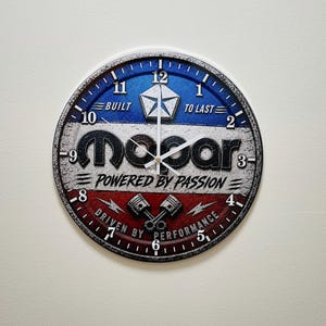 May include: A round wall clock featuring a Mopar logo with the text BUILT TO LAST, POWERED BY PASSION, and DRIVEN BY PERFORMANCE. The clock face has a blue, white, and red background with a pentastar emblem, two crossed pistons, and black numbers.