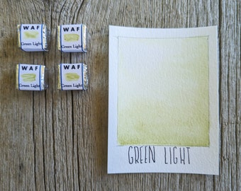 May include: Four small, silver-wrapped watercolor paint cubes labeled "WAF Green Light" are arranged near a watercolor swatch. The swatch is a rectangle of light green paint with the words "GREEN LIGHT" below it.