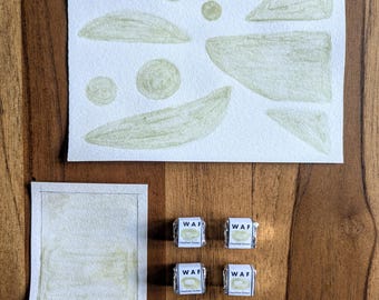 May include: A watercolor painting with abstract shapes in shades of green on white paper. Below, a smaller painting labeled "OWYHEE GREEN" and four wrapped candies are visible on a wooden surface.