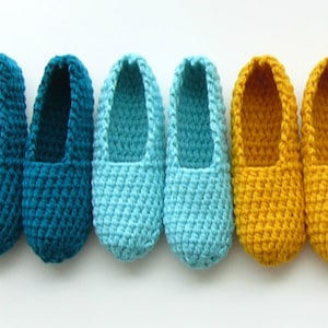 Op de afbeelding: Vijf paar gehaakte pantoffels in tinten van teal, blauw en geel. De pantoffels zijn in een rij van donkerst naar lichtst gerangschikt.
