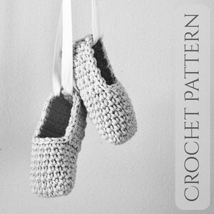 May include: Two gray crochet ballet slippers hanging from white ribbons. The slippers are made of a textured yarn and have a simple, classic design. The text "CROCHET PATTERN" is printed vertically on the right side of the image.