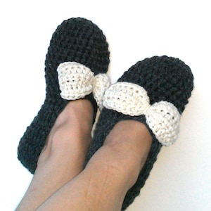 Può includere: Un paio di pantofole in crochet nero con fiocchi in crochet bianco. Le pantofole sono indossate ai piedi di una persona.