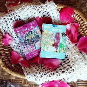 May include: Two sets of inspirational cards with colorful artwork and text. The first set is called "the Heart of Life" and features a dragonfly and the words "Authentic, Inner Compass, Trust". The second set is called "Soul Songs" and features a tree with purple flowers and the words "Gratitude, Celebration, Soft Smile, Leaving to the Sweet".