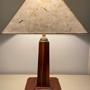 May include: A wooden table lamp with a square base and a tall, rectangular column. The lamp has a beige lampshade with a triangular shape and a textured surface.