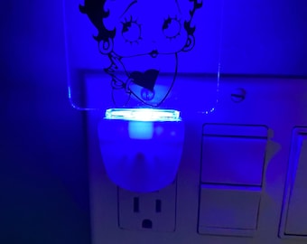 May include: A plug-in night light featuring a clear acrylic square panel with a black line art illustration of the cartoon character Betty Boop. The device emits a bright blue glow from its base, illuminating the character design against the wall.