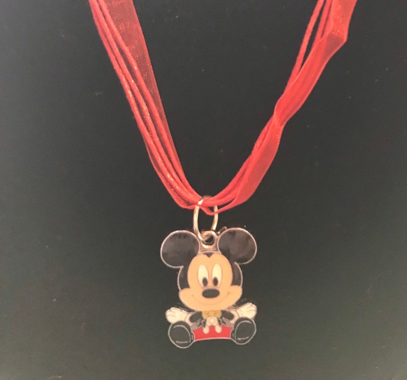 K&ouml;nnte beinhalten: Eine Halskette mit einem Mickey Mouse-Anh&auml;nger. Der Anh&auml;nger zeigt eine sitzende Mickey Mouse mit schwarzen Ohren, einer gelben Fliege und roten Shorts. Die Halskette hat ein rotes Band.