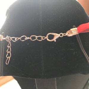 K&ouml;nnte beinhalten: Eine rote Bandkette mit einer silberfarbenen Kette und einem Verschluss. Die Halskette wird auf einer schwarzen Schaufensterpuppe pr&auml;sentiert. Das Band ist ein transparentes Material und die Kette ist in der L&auml;nge verstellbar.