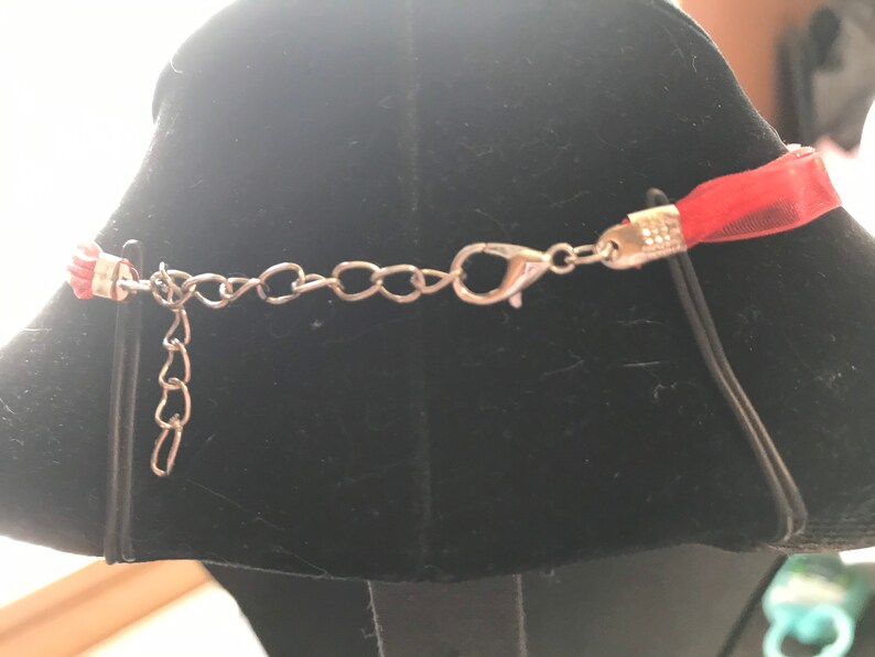 K&ouml;nnte beinhalten: Eine rote Bandkette mit einer silberfarbenen Kette und einem Verschluss. Die Halskette wird auf einer schwarzen Schaufensterpuppe pr&auml;sentiert. Das Band ist ein transparentes Material und die Kette ist in der L&auml;nge verstellbar.