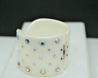 May include: A white plastic bangle bracelet with a hinged closure. The bracelet is decorated with iridescent rhinestones.