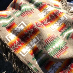 May include: A tan blanket with a Southwestern geometric pattern in red, orange, green, and white. The blanket has a fringed edge and a soft, textured appearance. The design features repeating diamond and rectangular shapes.