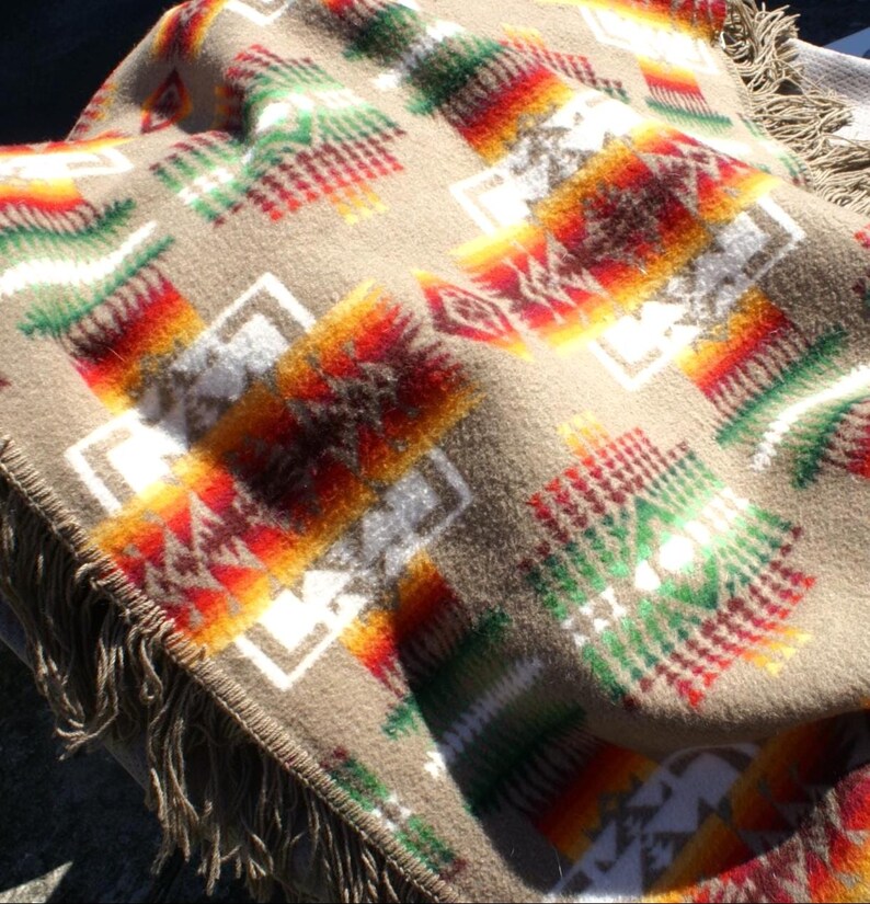 May include: A tan blanket with a Southwestern geometric pattern in red, orange, green, and white. The blanket has a fringed edge and a soft, textured appearance. The design features repeating diamond and rectangular shapes.