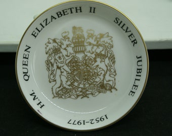 May include: White ceramic plate with a gold rim and a gold design of the royal coat of arms. The text "H.M. QUEEN ELIZABETH II SILVER JUBILEE 1952-1977" is printed around the edge of the plate.