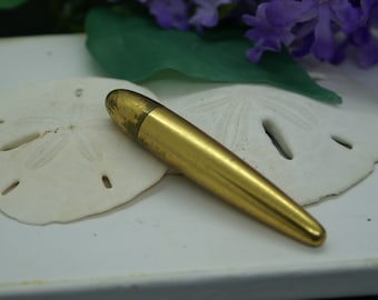 May include: A gold-colored metal lipstick tube with a rounded end. The tube is lying on a white surface.