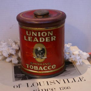 Peut inclure: Une boîte cylindrique en métal rouge avec un couvercle, portant l'inscription UNION LEADER SMOKING TOBACCO en lettres dorées. Un emblème ovale au centre affiche une illustration d'aigle noir. Le récipient a des bordures dorées en haut et en bas.