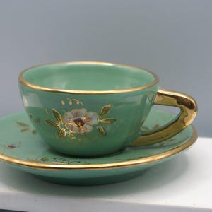 May include: A green teacup and saucer set with gold trim and floral designs. The cup has a gold handle and the saucer has a gold rim.