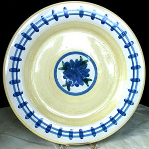May include: A round ceramic plate with a cream-colored base and a hand-painted blue floral design in the center. The rim features a decorative pattern of two concentric blue circles connected by short, evenly spaced vertical blue lines.