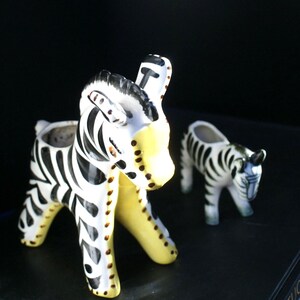 May include: Two ceramic zebra-shaped planters featuring a white base with black stripes and yellow accents on the legs and face. The larger zebra is in the foreground, while a smaller zebra planter is positioned behind it to the right.